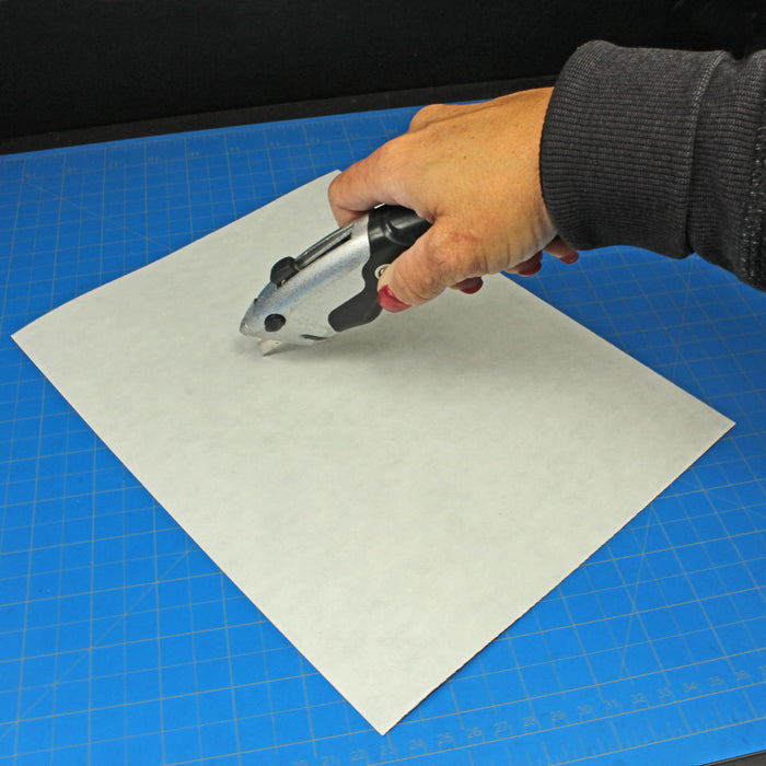 A person using a heavy-duty utility knife to cut a custom shape out of a  flexible magnetic sheet on a blue hobby cutting mat.