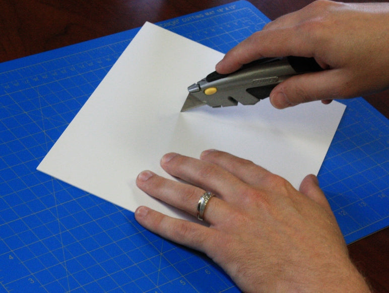 A hobby knife scoring a white styrene sheet to demonstrate the clean score and snap method for precise plasticard cutting.